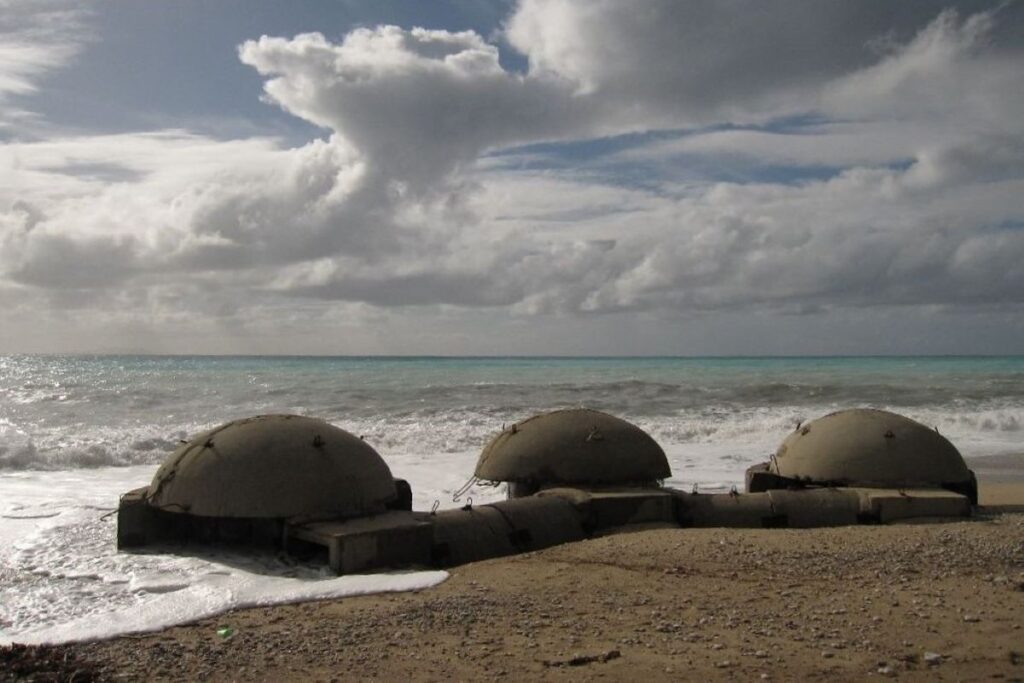 Three dome-shaped bunkers on a beach with waves and a cloudy sky.