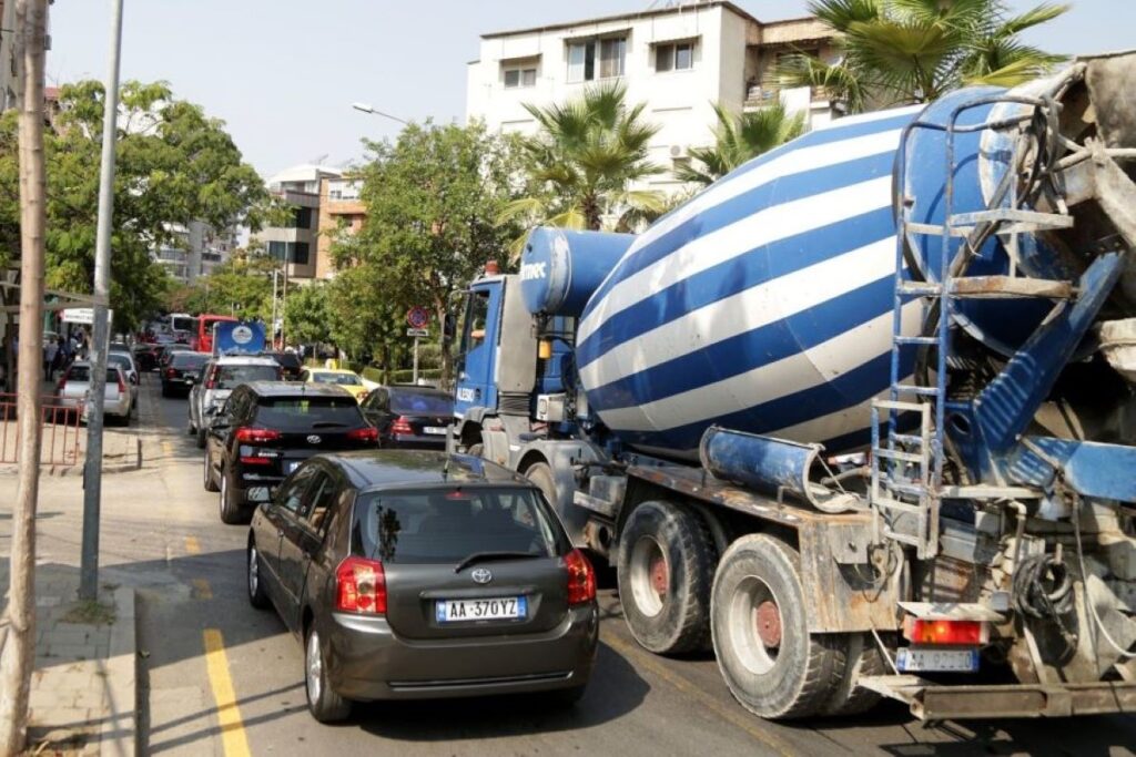 Traffic in Albania with cars, a cement truck, and pedestrians on a narrow urban street.