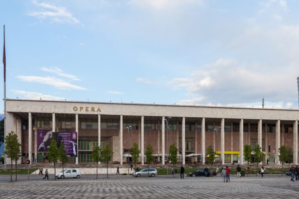 A large opera building with white columns, "OPERA" in gold letters, a modern high-rise in the background, and a plaza with trees and people in the foreground