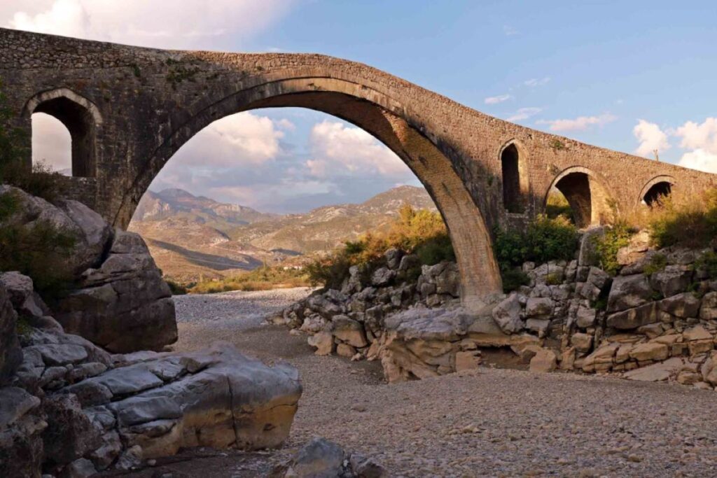 Historic Bridge of Mes with stone arches over a dry riverbed, set against rugged rocks and mountains