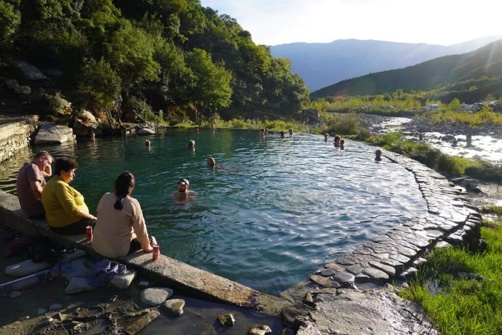 Benja Thermal Baths in Përmet, Albania, with visitors relaxing in a natural hot spring surrounded by lush greenery and mountains.