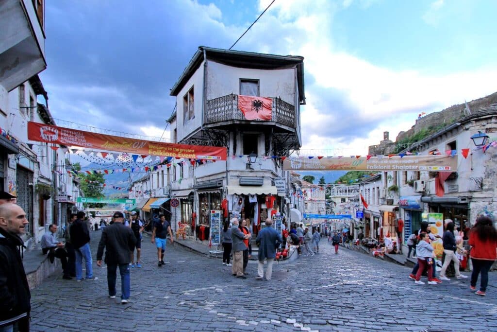 A bustling street in Gjirokastra, Albania, with traditional houses, shops, and decorations for a folklore event.